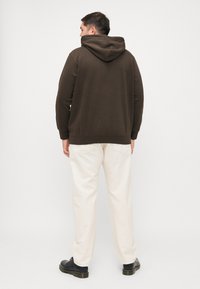 Sweatshirt à capuche marron avec une texture douce, associé à un pantalon couleur crème. L'ensemble est complété par des chaussures noires au design simple.