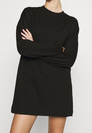 Persoon draagt een loszittende zwarte jurk met lange mouwen, met armen gekruist, staand tegen een eenvoudige lichte achtergrond.