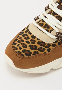 Braune und tanfarbene Sneaker mit einem Obermaterial im Leopardenmuster, aus Wildleder, flachen weißen Schnürsenkeln und einer strukturierten Sohle. Akzente setzen die Nähdetails.