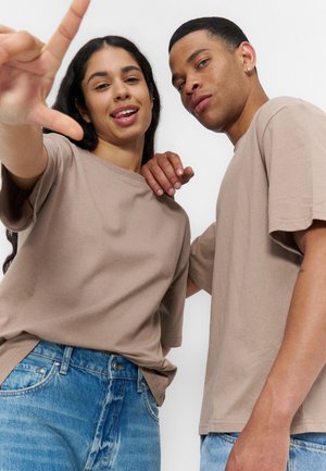 Deux jeunes adultes portant des chemises beige et des jeans, l’un tend la main vers l’avant, tous deux regardent l’appareil photo sur un fond blanc.
