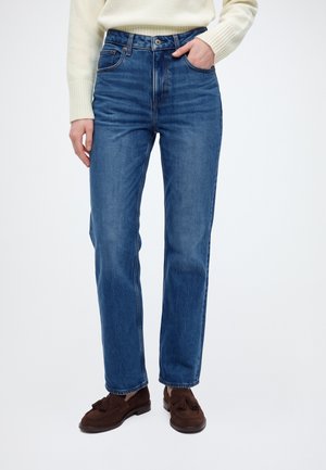 Un modèle cadré partiellement porte un jean bleu à jambes droites, un pull couleur crème et des mocassins marron à pompons. Une main est dans une poche, sur fond blanc.