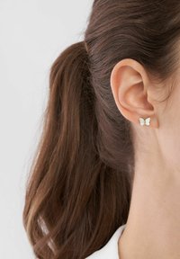 Orecchino a forma di farfalla in argento con finitura iridescente. Le ali texturizzate aggiungono dimensione. Indossato sul lobo di un orecchio di una persona con i capelli lunghi.