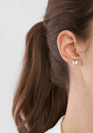 Orecchino a forma di farfalla in argento con finitura iridescente. Le ali texturizzate aggiungono dimensione. Indossato sul lobo di un orecchio di una persona con i capelli lunghi.