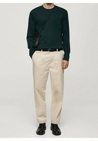 Homme portant une chemise à manches longues vert foncé, un pantalon beige, une ceinture noire, des bottes noires et un petit sac bandoulière noir, debout les mains dans les poches.