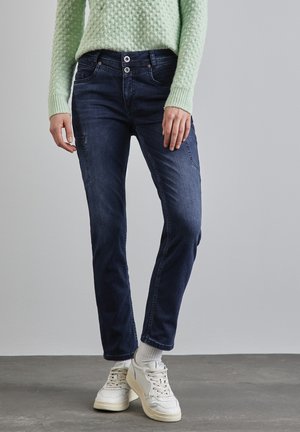 Jean bleu foncé à coupe slim, avec deux boutons à la taille et des poches latérales, associés à des baskets blanches et un pull texturé vert clair.