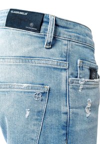 Jean en denim bleu clair avec un aspect usé. Il présente un patch logo noir à la ceinture et un détail de logo gris sur la poche arrière.