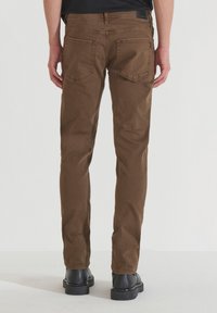 Pantalones ajustados marrones, tejido de algodón, cinco bolsillos con detalles de costura, y botas de cuero negras hasta el tobillo. Vista desde atrás.