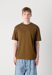 Braunes T-Shirt mit kurzen Ärmeln und Rundhalsausschnitt, versehen mit einem dezenten Logo auf der Brust, kombiniert mit hellblauen Jeans.