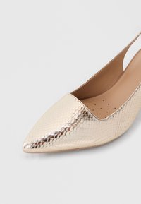Χρυσό μεταλλιζέ παπούτσι slingback με μυτερή μύτη, υφή, λεία καφετί εσωτερική επένδυση και μικρές οπές κοντά στη φτέρνα.