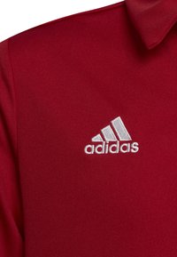 Polo di colore rosso realizzato in tessuto testurizzato, con un logo Adidas ricamato di colore bianco nella zona sinistra del petto. Design con colletto corto.