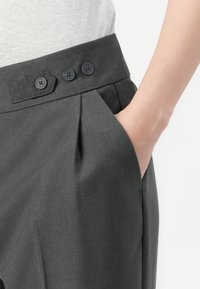 Person wearing dark gray pants with three black buttons on the waistband and hand placed inside the right pocket.
