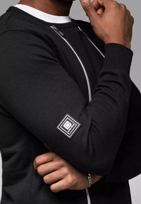 Sudadera negra con cremallera y cuello blanco, con mangas texturizadas y un parche cuadrado de logo en el brazo. Material de algodón suave.