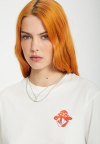 White t-shirt featuring a colorful graphic of a mushroom and diamond design on the chest. Two layered silver chain necklaces.