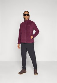 Giacca in pile viola con chiusura a zip e tasche laterali, abbinata a pantaloni neri e stivaletti marroni, su uno sfondo semplice.