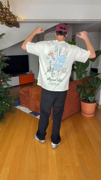 Light-colored t-shirt with a graphic of a blue chair and table, paired with black pants and silver shoes, set in a home environment.