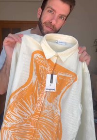 Balta tekstūruota marškiniai su oranžiniu abstrakčiu dizainu. Yra apykaklė ir mygtukų uždarymas, prekės ženklo etiketė matoma prie kaklo.