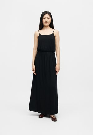 Γυναίκα με μακριά σκούρα μαλλιά, φορώντας ένα μακρύ μαύρο φόρεμα χωρίς μανίκια και σκούρα καφέ μπαλαρίνες, στέκεται μπροστά σε απλό φωτεινό φόντο.