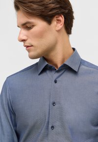 Een button-up shirt in een licht marineblauwe kleur, met een subtiele textuur, een puntige kraag en donkere knopen aan de voorkant.