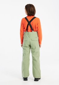 Groene sneeuwbroeken met verstelbare zwarte bretels, rechte pijpen en twee achterzakken, gedragen over een oranje lange mouwen top.
