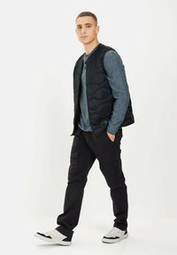 Schwarze gesteppte Weste über einem blauen, gestreiften Langarmhemd, kombiniert mit schwarzen Cargo-Hosen und weißen Sneakers. Schlichtes, funktionales Design.
