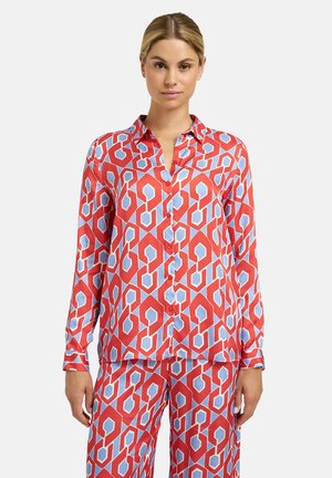 Frau trägt langärmliges Hemd mit Knöpfen und passende Hose mit rotem und hellblauem geometrischem Muster, steht vor schlichtem Hintergrund.