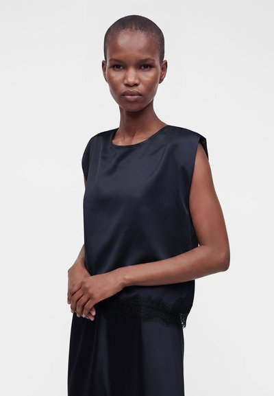 Femme aux cheveux courts portant un haut en satin noir sans manches avec bordure en dentelle, debout les mains jointes devant un fond clair uni.