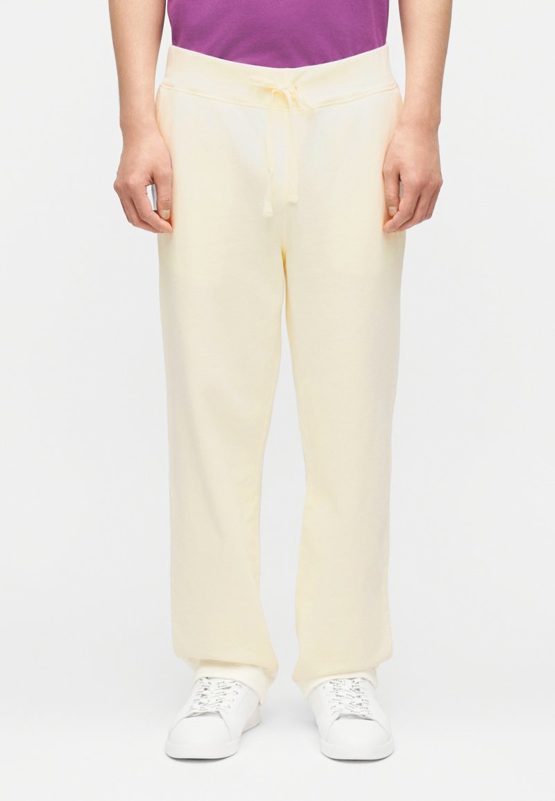 Άτομο που φοράει κρεμ φόρμες με κορδόνι και λευκ ά αθλητικά παπούτσια, στέκεται μπροστά σε απλό φωτεινό φόντο.