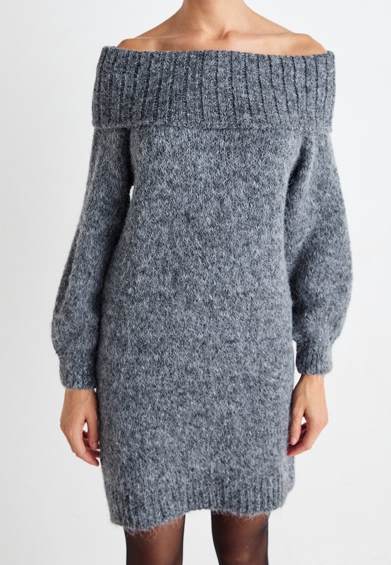 Femme portant une longue robe pull grise à épaules dénudées, manches longues, avec poignets et col côtelés.