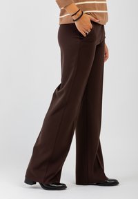 Persona che indossa pantaloni larghi marroni con tasche, scarpe nere con tacco basso e un maglione marrone con strisce bianche, in piedi davanti a uno sfondo semplice.