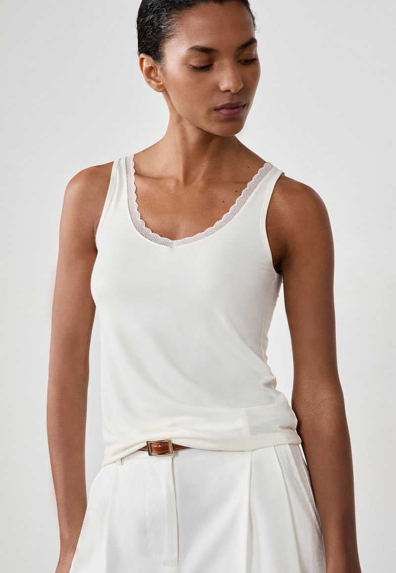 Donna che indossa un top bianco senza maniche con scollatura in pizzo e pantaloni bianchi a vita alta con cintura marrone, che guarda verso il basso a sinistra.