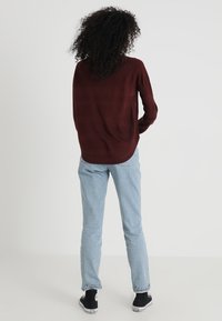 Maroon sweater with a textured pattern, long sleeves, and a curved hem. Paired with light blue jeans and black sneakers.