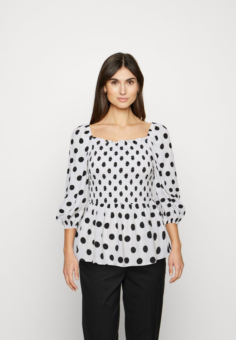 Blusa branca com bolinhas pretas, decote quadrado e corpete franzido. Apresenta mangas bufantes e um detalhe em bainha evasé.