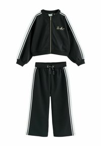 Čierny tréningový oblek s bomber bundou na zips a širokými nohavicami. Obsahuje biele pruhy na rukávoch a nohách, vyrobené z hladkej tkaniny.