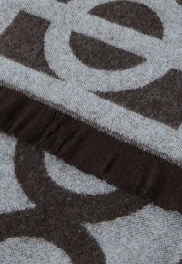 Wełniany koc w wzory w kolorach szarym i ciemnobrązowym, z frędzlami na krawędziach oraz abstrakcyjnymi kształtami w kontrastowych odcieniach.