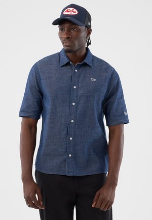 Uomo con camicia blu navy a maniche corte con bottoni e cappellino blu navy con logo rosso e bianco, in piedi davanti a uno sfondo semplice.