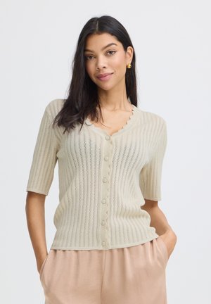 Žena s tmavými vlasmi, oblečená v béžovom krátkhorukávovom zapínacom kardigáne a svetlohnedých nohaviciach, stojaca s rukami v kapse pred jednoduchým pozadím.