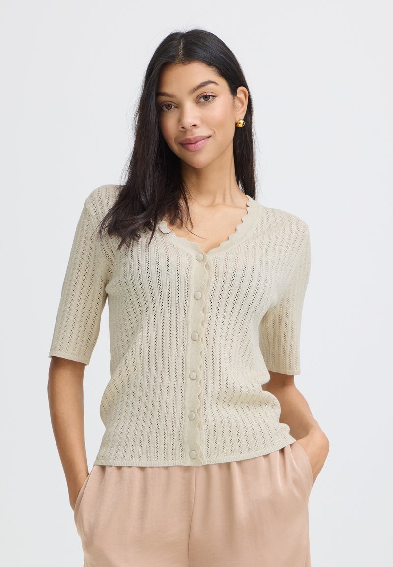 Femme aux cheveux foncés portant un cardigan beige à manches courtes boutonné et un pantalon beige clair, debout les mains dans les poches devant un fond uni.