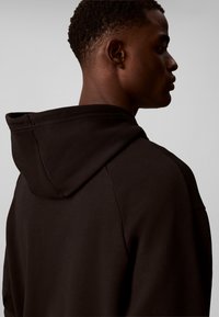 Jeune homme aux cheveux courts et bouclés portant un sweat à capuche noir, vu de dos et de profil sur un fond uni.