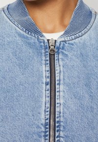 Svetle modrá džínová bombera s žebrovaným límcem a manžetami, s centrálním zipem a kontrastními švy.