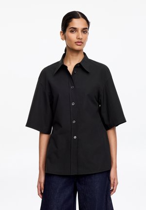 Kobieta o ciemnych włosach związanych z tyłu, ubrana w czarną koszulę na krótki rękaw z guzikami oraz ciemnoniebieskie szerokie spodnie, stoi na jednolitym białym tle.