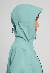 Veste imperméable bleu clair avec capuche ajustée, cordons élastiques et zips contrastés. Texture lisse, design minimaliste.