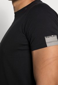 T-shirt en coton noir à manches courtes, avec une bande grise et le logo "REPLAY" sur la manche gauche. Coupe ajustée au corps.