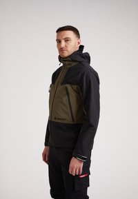 Giacca nera e verde oliva con cappuccio regolabile, chiusura frontale con zip, due tasche sul petto e un logo discreto sul petto. Tessuto testurizzato.