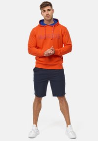Orangene Kapuzenjacke mit blauer Innenseite, ausgestattet mit gedrucktem Text. Kombiniert mit dunklen Shorts und weißen Sneakern. Fokus auf lässigem Sportbekleidungsstil.