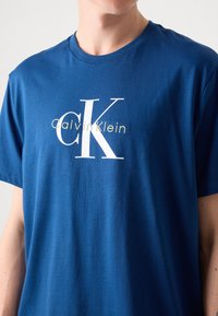 Camiseta de algodón azul con cuello redondo, que presenta el logo blanco "CK" y el texto "Calvin Klein" en amarillo debajo. Manga corta, corte relajado.