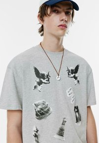 Grå bomuld t-shirt med sorte grafiske tryk af statuer og engle, rund halsudskæring, korte ærmer og en brun snor halskæde med et vedhæng.