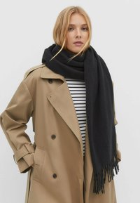 Sort uldhalstørklæde med frynser, båret over en stribet skjorte og en beige trench coat med store knapper og en afslappet pasform.