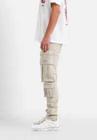 Beige cargobroek met meerdere zijzakken, ritsen en een slank ontwerp, gecombineerd met witte sportieve schoenen en een graphic t-shirt.