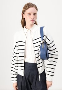 Hvit strikket cardigan med svarte striper, glidelås og ribbede mansjetter. Brukt over en hvit skjorte med krage, kombinert med mørke bukser og en blå veske.