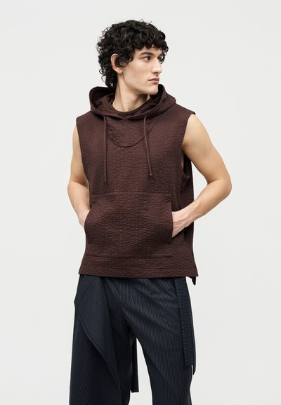 Jonge man met krullend haar, gekleed in een mouwloze bruine hoodie met structuur en losse donkere broek met fijne strepen, kijkt naar rechts met zijn handen in zijn zakken.